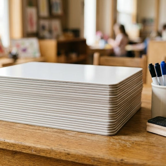 A stack of thin frameless DryErase student lap boards on a classroom table beside a cup of dry erase markers and wood-block erasers